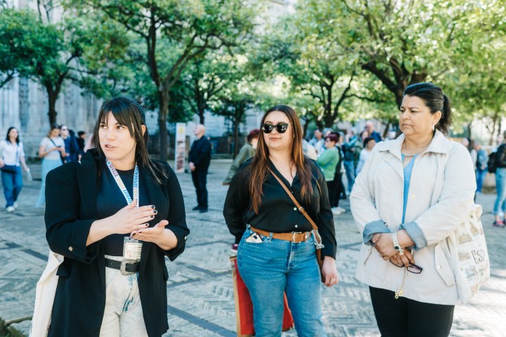 Three women standing outdoors, one speaking, in a leafy courtyard.