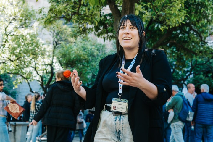 Woman speaking outdoors, wearing a lanyard, with trees and people in the background.