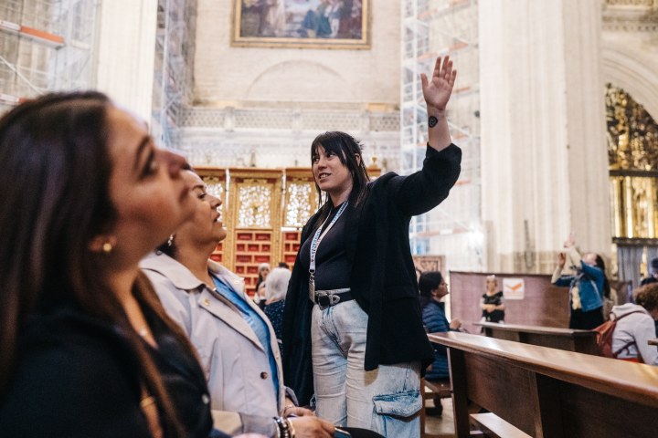 A tour guide gestures upward in a cathedral while speaking to visitors.