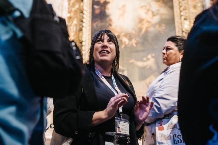 Woman with lanyard speaks to group indoors near artwork, people around her.