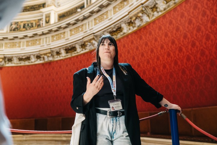 Person speaking in ornate room with red walls, wearing lanyard and ID badge.