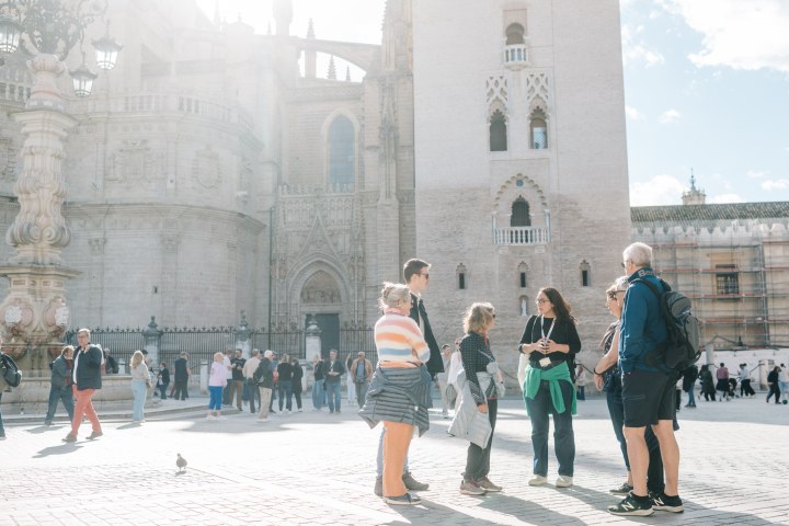 Tour group stands in sunny plaza with historic building in background.