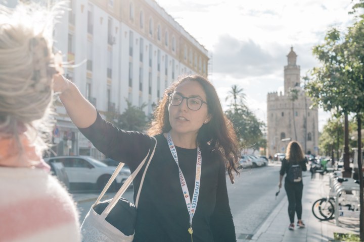 Woman with glasses gestures while speaking on a sunny street near a historical tower.