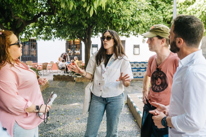 Four people talking outdoors under green trees on a stone path.