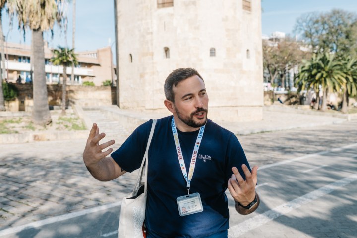 Man in blue shirt speaking animatedly outside near a historical tower.