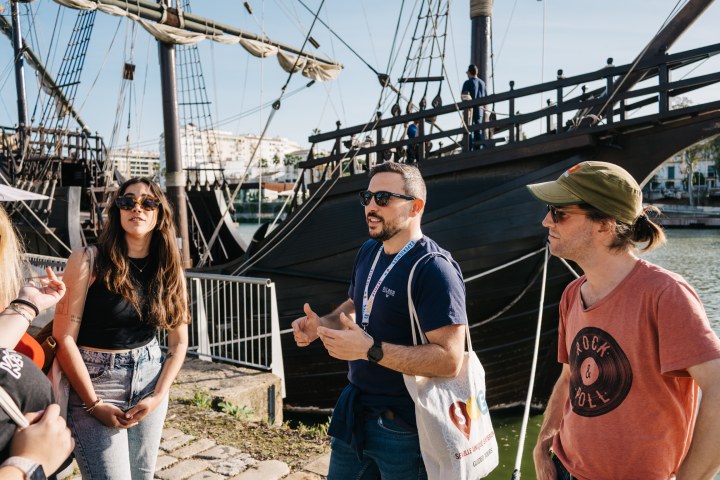 Three people talking near a wooden ship docked by a riverbank under a clear sky.
