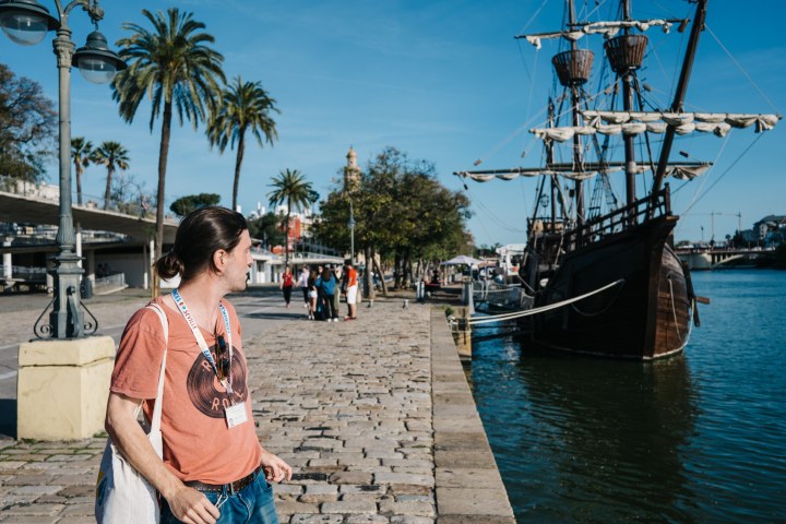 Person on a riverside promenade near a docked tall ship and palm trees under a clear blue sky.
