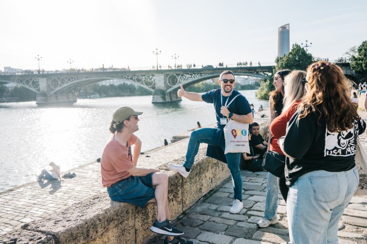 Group of people laughing by a river with a bridge in the background.