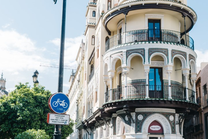 People standing near ornate building with balconies and round tower on a sunny day.