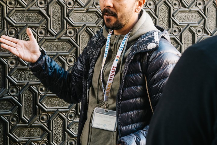 Man with lanyard gesturing in front of ornate patterned wall.