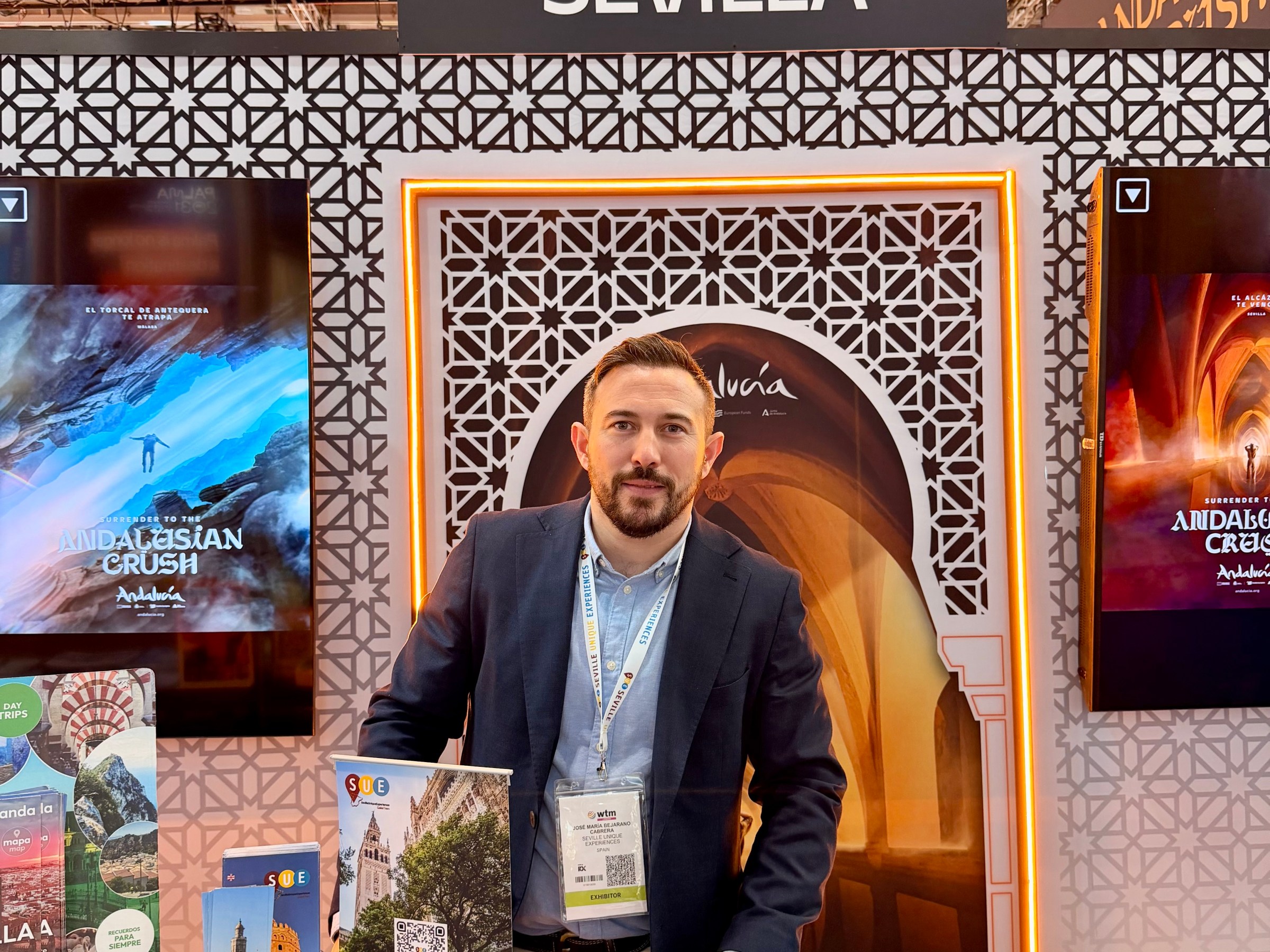 Man in a suit stands at a promotional booth for Sevilla, surrounded by brochures.