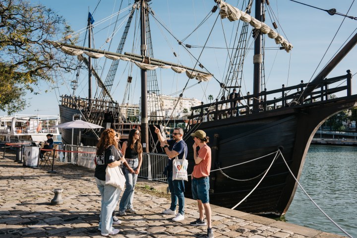 Tourists stand near a historic wooden ship docked on a sunny waterfront.
