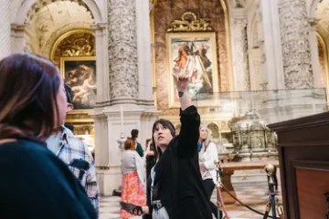 A person gestures upward in a museum with ornate paintings and arches in the background.
