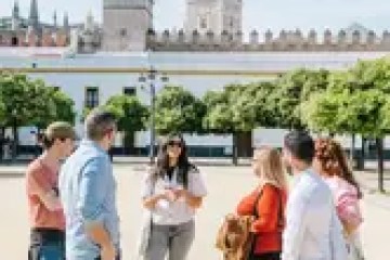People in a courtyard near a historic tower and trees on a sunny day.