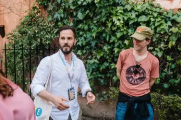 Two men talking outside, one in a white shirt, the other in a red shirt and cap, with ivy in the background.