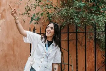 Person smiling and pointing upwards near a fence with vines.