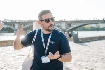 Man in sunglasses gesturing near a river with a bridge in the background.