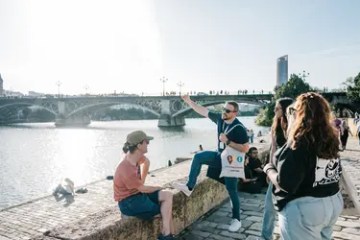 People sitting and talking by a riverbank with a bridge in the background.