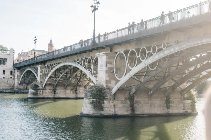 Stone bridge with ornate railings over a calm river, people walking on top.