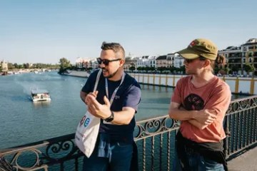 Two men talking on a riverside bridge with a boat in the water.